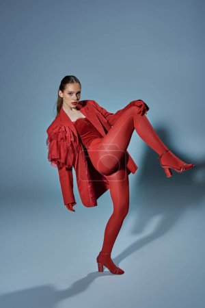 stylish model in red outfit looking at camera while posing with raised leg on grey background
