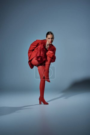 fashionable model in red outfit looking at camera while posing with raised leg on grey background