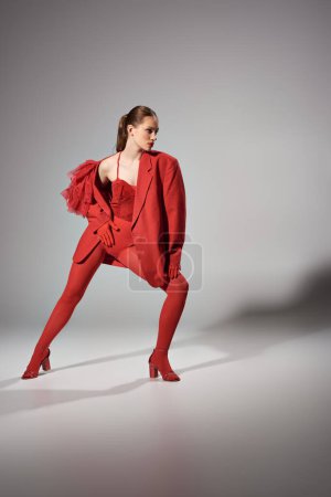 stylish woman in red outfit looking at camera while posing on grey background, fashion photography