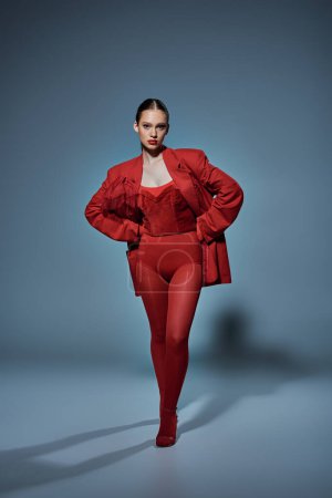 young woman in red outfit looking at camera while posing with hands on hips on grey background