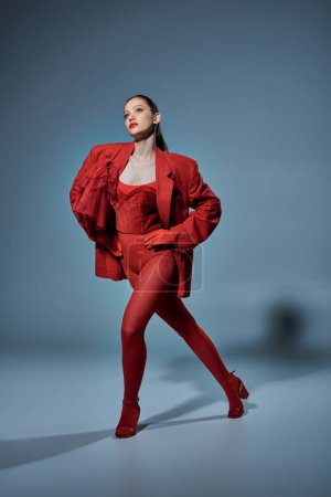 young model in red outfit looking at camera while posing with hands on hips on grey background