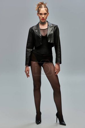 stylish young woman with pearls in hair posing in leather jacket and black tights on grey background