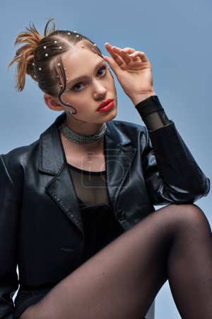 young trendy model in leather jacket and black tights looking at camera on grey backdrop, fashion
