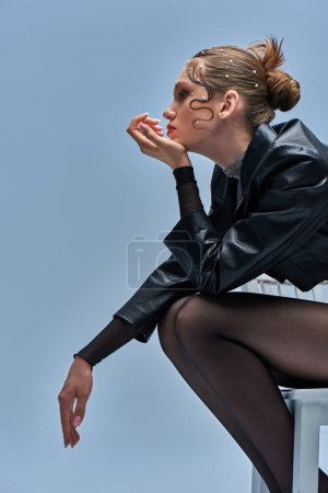 Nachdenkliche junge Frau in schwarzer Lederjacke und Strumpfhose auf Stuhl vor grauem Hintergrund sitzend, Perlenketten
