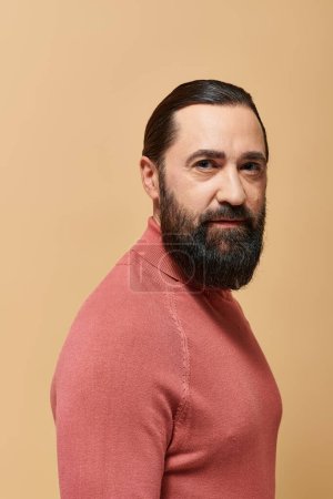 Porträt, ernster, gutaussehender Mann mit Bart posiert in rosa Rollkragenpullover auf beigem Hintergrund