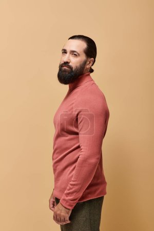 portrait, bel homme sérieux avec barbe posant en pull col roulé rose sur fond beige