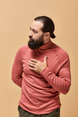 portrait of serious and handsome man with beard posing in pink turtleneck jumper on beige background t-shirt #684013116