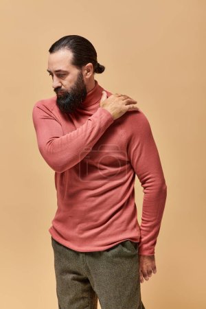 retrato, hombre serio y guapo con barba posando en jersey de cuello alto rosa sobre fondo beige