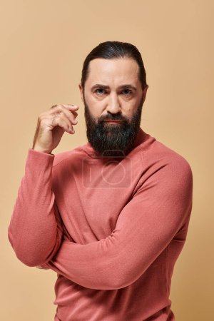 ernster und gutaussehender Mann mit Bart posiert in rosa Rollkragenpullover auf beigem Hintergrund, Portrait