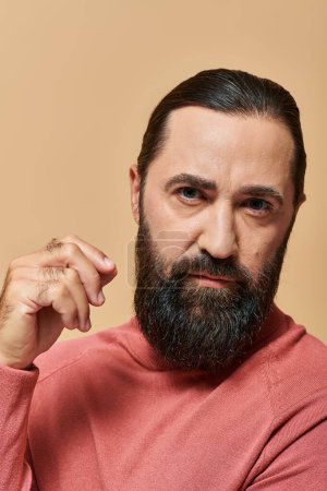 handsome serious man with beard posing in pink turtleneck jumper on beige background, portrait