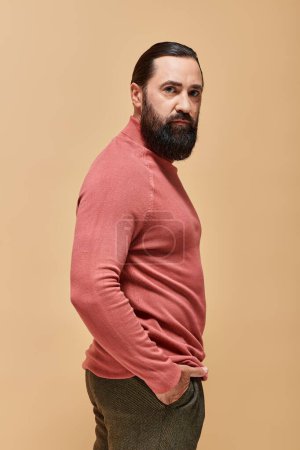 good looking and serious man with beard posing in pink turtleneck jumper on beige, portrait