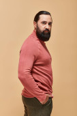 good looking and serious man with beard posing in pink turtleneck jumper on beige, portrait Poster #684013482