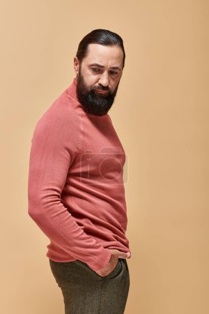 retrato, modelo serio y guapo con barba posando en jersey de cuello alto rosa sobre fondo beige