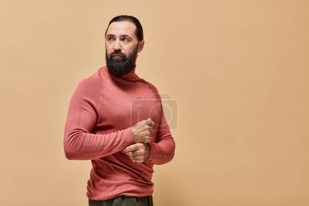 hombre guapo y serio con barba posando en jersey de cuello alto rosa sobre fondo beige, retrato