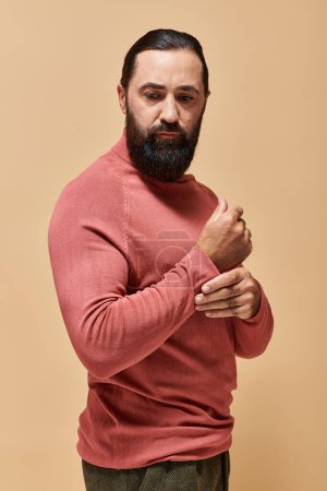 seriöser gutaussehender Mann mit Bart posiert in rosa Rollkragenpullover auf beigem Hintergrund, Portrait