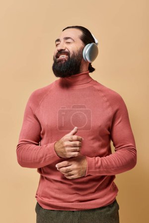 happy bearded man in turtleneck jumper listening music in wireless headphones, smiling on beige