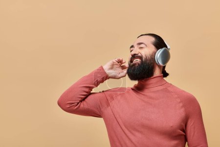 cheerful bearded man in turtleneck jumper listening music in wireless headphones, smiling on beige