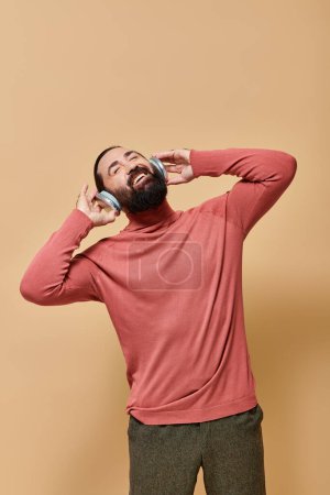 bearded and happy man in turtleneck jumper listening music in wireless headphones, beige backdrop