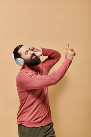 bearded and cheerful man in turtleneck jumper listening music in wireless headphones, beige backdrop