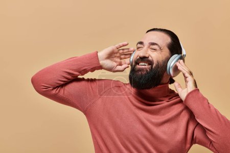 bearded and positive man in turtleneck jumper listening music in wireless headphones, beige backdrop