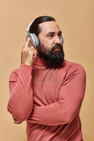 schöner bärtiger Mann im Rollkragenpullover, der Musik in drahtlosen Kopfhörern auf beigem Hintergrund hört