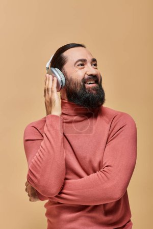 smiling and bearded man in turtleneck jumper listening music in wireless headphones, beige backdrop
