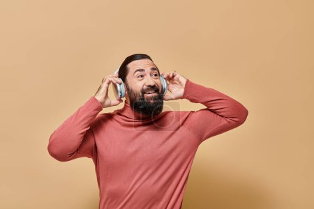 pleased bearded man in turtleneck jumper listening music in wireless headphones, beige backdrop