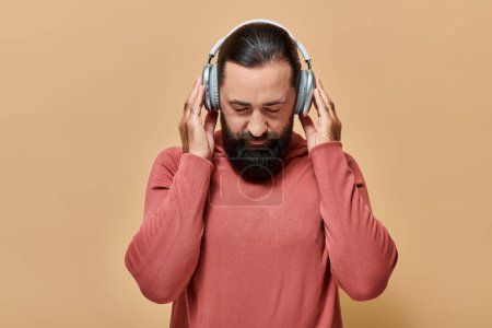 handsome bearded man in turtleneck jumper listening music in wireless headphones on beige background