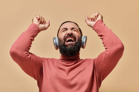 excited bearded man in turtleneck jumper listening music in wireless headphones, beige backdrop