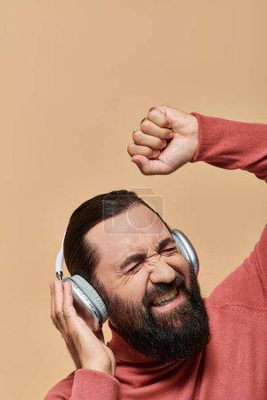 thrilled bearded man in turtleneck jumper listening music in wireless headphones, beige background