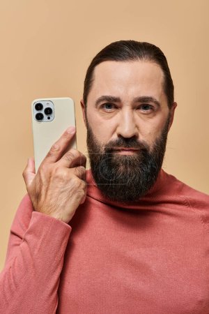portrait of good looking bearded man in turtleneck jumper holding smartphone on beige background