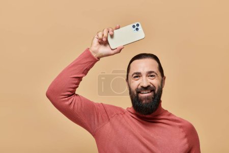 retrato de hombre barbudo feliz en jersey de cuello alto sosteniendo teléfono inteligente sobre fondo beige