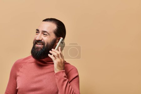 portrait of cheerful bearded man in turtleneck jumper talking on smartphone on beige background
