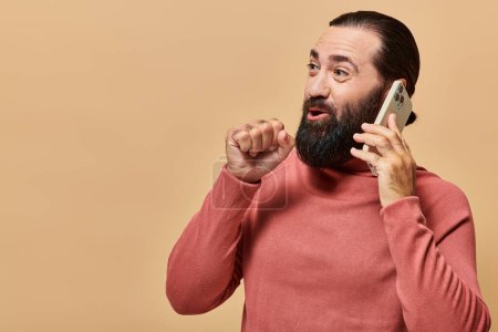 portrait of surprised bearded man in turtleneck jumper talking on smartphone on beige background