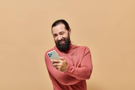 retrato de hombre barbudo alegre en jersey de cuello alto sosteniendo teléfono inteligente sobre fondo beige
