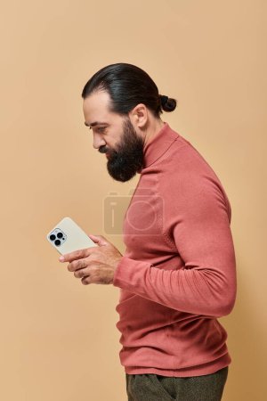 portrait of bearded good looking man in turtleneck jumper holding smartphone on beige background