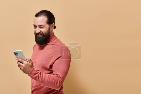portrait d'homme barbu gai en pull à col roulé tenant smartphone sur fond beige