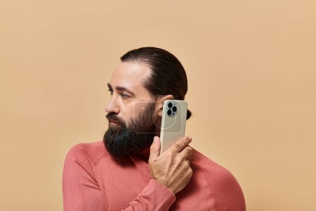 portrait of good looking bearded man in turtleneck jumper holding smartphone on beige background