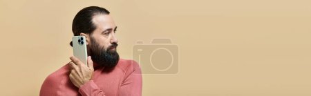 portrait of good looking bearded man in turtleneck jumper holding smartphone on beige, banner