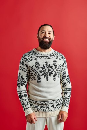 glücklicher bärtiger Mann im Winterpullover mit Ornament lächelnd auf rotem Hintergrund, Frohe Weihnachten