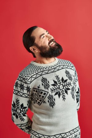 Foto de Hombre barbudo feliz en suéter de invierno con adorno sonriendo en el telón de fondo rojo, Feliz Navidad - Imagen libre de derechos