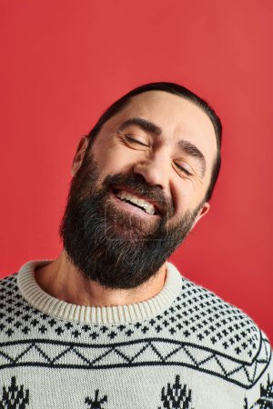 Photo for Happy bearded man in winter sweater with ornament smiling on red backdrop, Merry Christmas - Royalty Free Image
