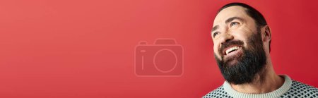 happy bearded man in winter sweater with ornament smiling on red backdrop, Merry Christmas banner