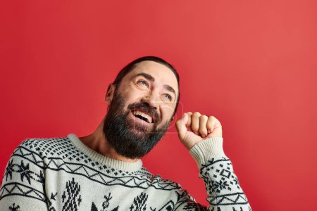 cheerful bearded man in winter sweater with ornament knocking on red backdrop, Merry Christmas