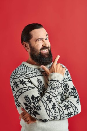 cheerful bearded man in winter sweater with ornament pointing on red backdrop, Merry Christmas