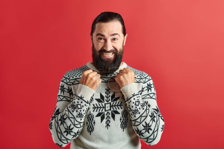 hombre barbudo excitado en suéter de invierno con adorno sonriendo en el telón de fondo rojo, Feliz Navidad