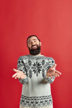 happy bearded man in winter sweater with ornament gesturing on red backdrop, Merry Christmas