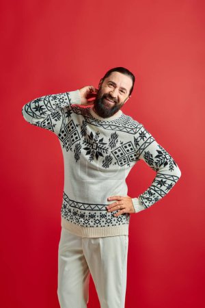 happy bearded man in winter sweater posing with hand on hip on red backdrop, Merry Christmas
