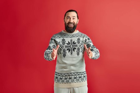 positiver bärtiger Mann im Weihnachtspullover mit Ornament gestikulierend auf rotem Hintergrund, Winterurlaub
