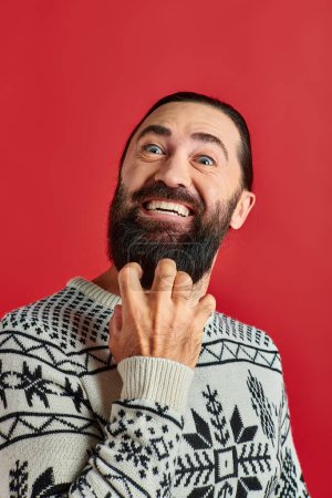 hombre barbudo apasionado en suéter de Navidad con adorno sonriendo en el telón de fondo rojo, vacaciones de invierno